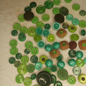 May include: A collection of various green buttons in different shades and sizes. The buttons are round, with two or four holes, and some have decorative patterns. The color palette ranges from dark forest green to light lime green.