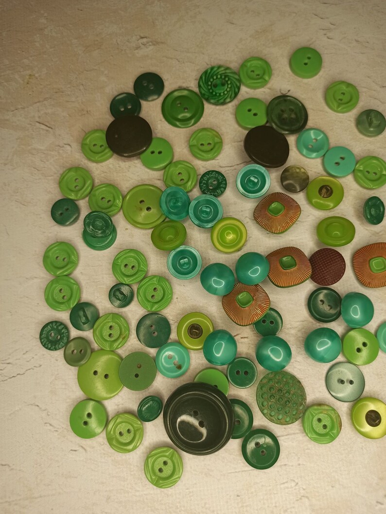 May include: A collection of various green buttons in different shades and sizes. The buttons are round, with two or four holes, and some have decorative patterns. The color palette ranges from dark forest green to light lime green.
