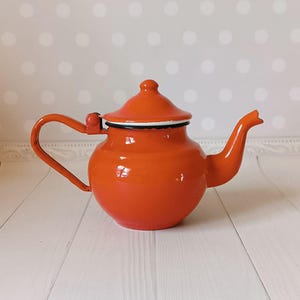 Puede incluir: Una tetera de esmalte naranja brillante con un borde blanco y un asa negra. La tetera tiene un cuerpo redondeado y un pequeño pico.