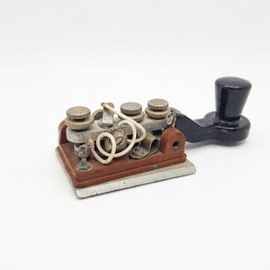 May include: A telegraph key featuring a brown rectangular base, a silver metal plate, and a black plastic knob. The device includes metallic adjustment screws, a small coiled white wire, and exposed electrical components mounted on the top surface.