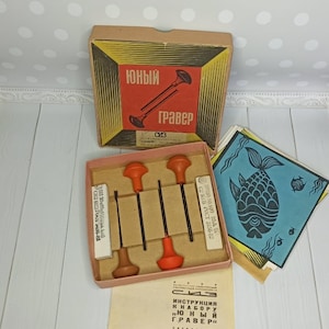 Może przedstawiać: Drewniana skrzynka vintage zawierająca zestaw narzędzi do grawerowania. Skrzynka jest oznaczona napisem "ЮНЫЙ ГРАВЕР" (Młody Grawer) i zawiera zestaw czterech narzędzi do grawerowania z czerwonymi i brązowymi uchwytami, mały blok białej gumki do ścierania oraz instrukcje w języku rosyjskim.