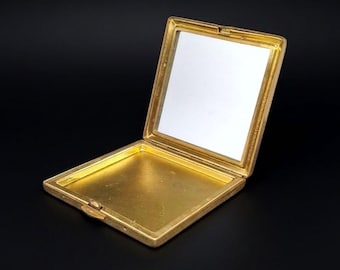 Könnte beinhalten: Ein quadratisches, goldfarbenes Metallpuderetui mit Klappdeckel. Das Innere verfügt über einen quadratischen Spiegel im oberen Bereich und ein leeres, glattes, goldfarbenes Bodenfach. Das Etui hat einen kleinen Verschlussmechanismus an der Vorderkante.