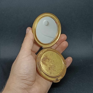 Può includere: Uno specchio compatto dorato con una superficie usurata. Lo specchio è aperto e l'interno è bianco. Il compatto ha una piccola iscrizione che dice "Made in USA".