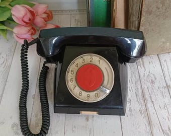 May include: A black rotary dial phone with a red button in the center of the dial. The phone has a black coiled cord.