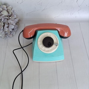 Puede incluir: Un teléfono de disco vintage con una base azul turquesa y un auricular rojo coral. El teléfono tiene un dial negro con números blancos.