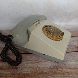 May include: A vintage rotary dial phone with a gray and white color scheme. The phone has a coiled cord and a rotary dial with a clear plastic cover.