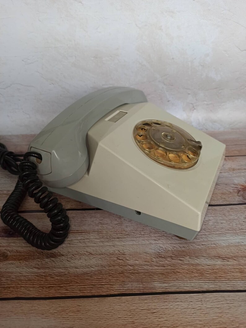 May include: A vintage rotary dial phone with a gray and white color scheme. The phone has a coiled cord and a rotary dial with a clear plastic cover.