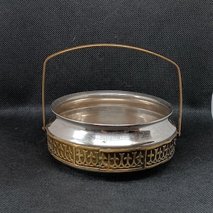 Puede incluir: Un cuenco de plata con un borde y un asa dorados. El cuenco tiene un patrón decorativo alrededor del borde.