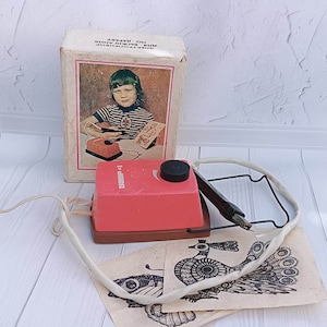 May include: A pink and black electric pen tool with a white cord and a black metal stand. The tool is on a wooden surface with three pieces of paper with black and white drawings of birds. The box for the tool is in the background and has a picture of a child using the tool. The box says "ЭЛЕКТРОГРАФ" and "АВТОР В.И.КНЯЗЕВА" and "ДО 12 ЛЕТ".