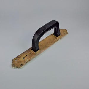 Puede incluir: Un tirador de puerta antiguo con un agarre negro y una placa trasera dorada y texturizada. La placa trasera presenta un ojo de cerradura y bordes decorativos. El tirador está diseñado para la funcionalidad y añade una estética vintage.