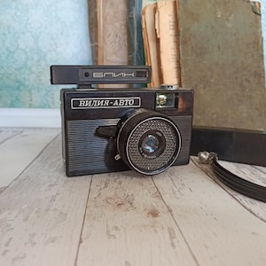 Puede incluir: Una cámara de película vintage negra con una lente y un accesorio de flash. La cámara tiene el texto "ВИЛИЯ-АВТО" y "БЛИК" en ella.