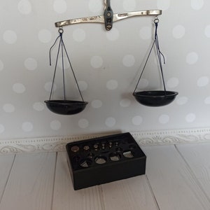 Puede incluir: Una balanza de equilibrio vintage de plata con dos cuencos negros colgando de ella. La balanza est&aacute; sobre una superficie blanca con una caja negra que contiene un juego de pesas.