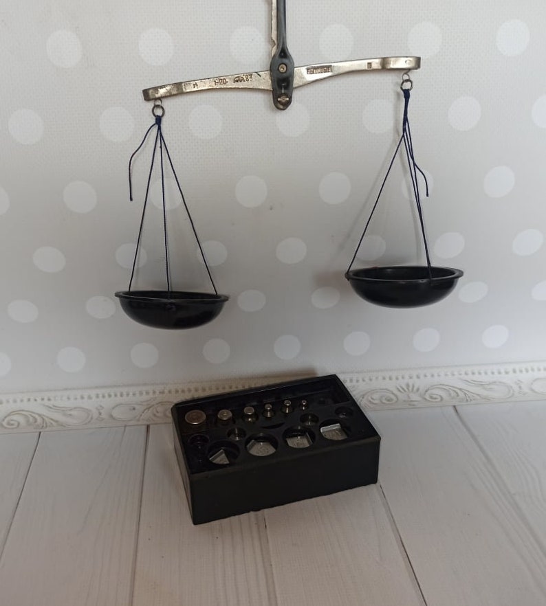 Puede incluir: Una balanza de equilibrio vintage de plata con dos cuencos negros colgando de ella. La balanza est&aacute; sobre una superficie blanca con una caja negra que contiene un juego de pesas.