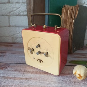 May include: A vintage red and cream-colored alarm clock with a gold handle and knobs. The clock has a square shape and is set against a backdrop of old books and a white brick wall. The clock's face features three knobs and a small, stylized symbol.