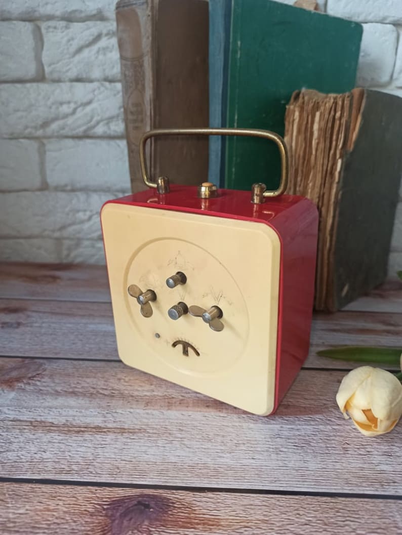 May include: A vintage red and cream-colored alarm clock with a gold handle and knobs. The clock has a square shape and is set against a backdrop of old books and a white brick wall. The clock's face features three knobs and a small, stylized symbol.