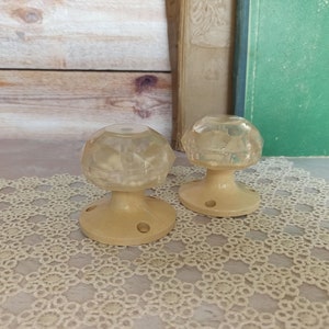 Op de afbeelding: Twee vintage deurknoppen met een helder, facetgeslepen glasontwerp en een crèmekleurige basis. De knoppen liggen op een wit gehaakt doily.