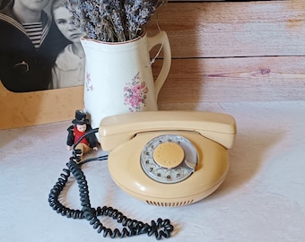 Pode incluir: Um telefone de disco rotativo vintage de cor amarelo claro com um cabo espiralado preto. O telefone tem um dial circular com números pretos e um botão central amarelo. O telefone está sobre uma superfície branca.