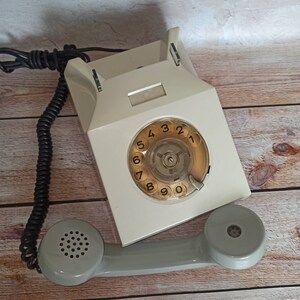 May include: A vintage rotary dial phone with a white body and a gray handset. The phone has a circular dial with black numbers 1 through 9 and 0. The phone is on a wooden surface.