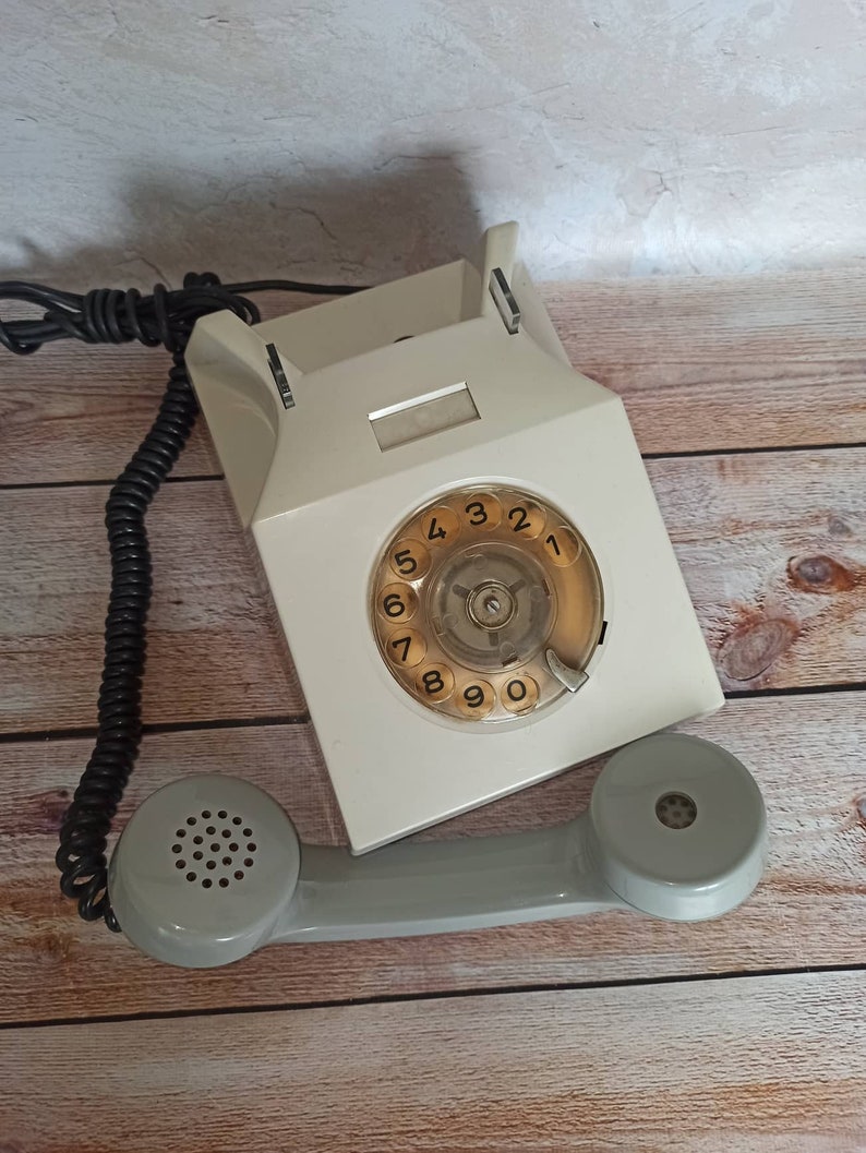 May include: A vintage rotary dial phone with a white body and a gray handset. The phone has a circular dial with black numbers 1 through 9 and 0. The phone is on a wooden surface.