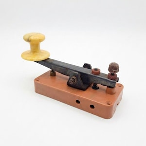 May include: An antique telegraph key. It features a brown base, a black lever, and a yellow knob. The key has two red adjustment screws and a brown knob. This vintage item is likely from the early 20th century.