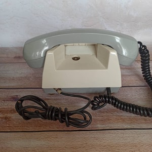 May include: A vintage rotary dial phone with a gray handset and a white base. The phone has a coiled cord and a separate black cord.