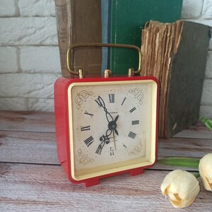 May include: A vintage red and gold alarm clock with a cream-colored face and black Roman numerals. The clock has a gold handle and is set against a backdrop of old books. The clock is approximately 4 inches tall.