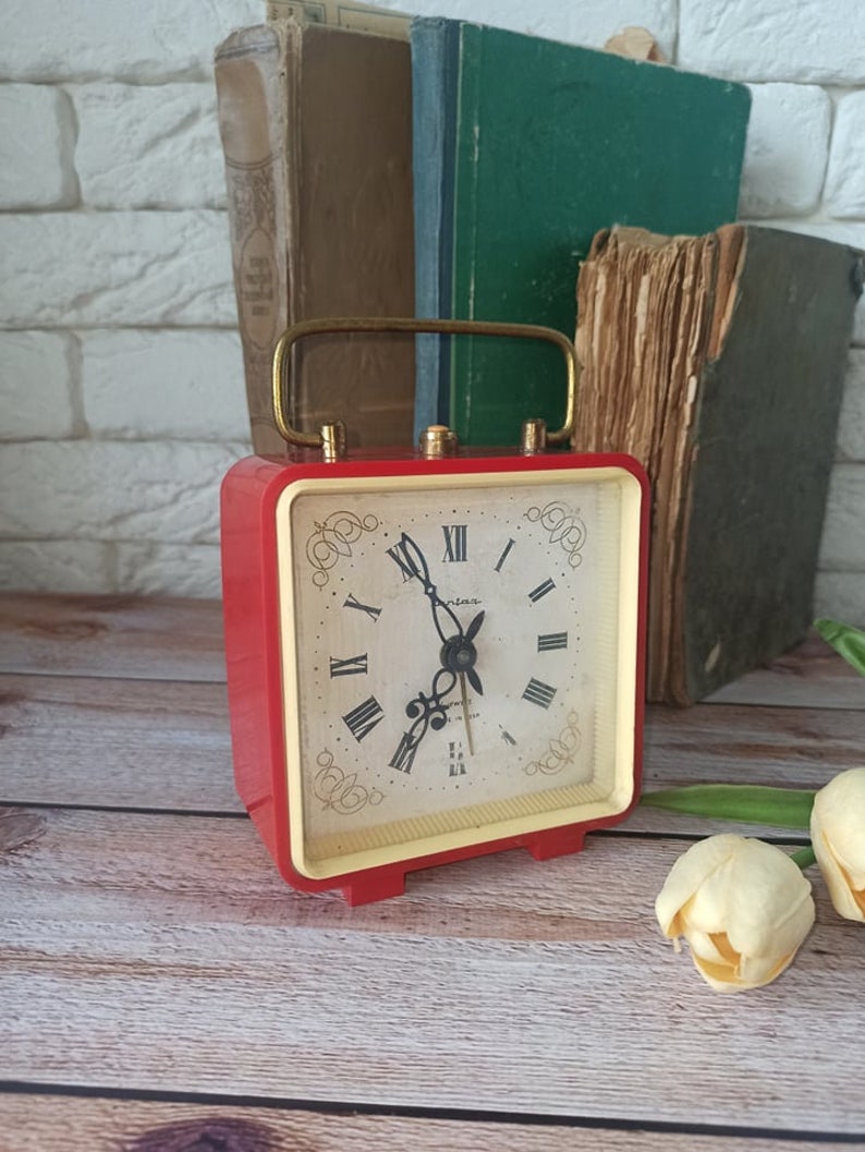 May include: A vintage red and gold alarm clock with a cream-colored face and black Roman numerals. The clock has a gold handle and is set against a backdrop of old books. The clock is approximately 4 inches tall.