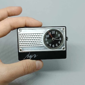 Könnte beinhalten: Ein kleines, schwarz-silbernes Taschenradio mit einem Zifferblatt. Das Zifferblatt hat schwarze Zahlen und Zeiger sowie einen roten Sekundenzeiger. Das Radio hat ein silbernes Lautsprechergitter und ein schwarzes Kunststoffgehäuse.