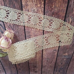 May include: A beige crocheted lace trim with a delicate pattern. The lace is displayed on a brown wooden background.