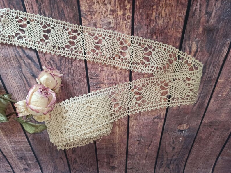 May include: A beige crocheted lace trim with a delicate pattern. The lace is displayed on a brown wooden background.