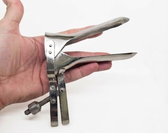 Może przedstawiać: Srebrzysty metalowy wziernik dopochwowy z dwoma łopatkami w kształcie dzioba kaczki, utrzymywanymi w pozycji otwartej za pomocą gwintowanego mechanizmu śruby regulacyjnej znajdującego się u podstawy uchwytu. Instrument medyczny ma gładkie wykończenie.