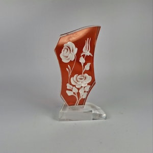 May include: A decorative, orange-red glass vase with a clear, crystal base. The vase features white, raised floral designs of roses and buds. The vase is approximately 6 inches tall.