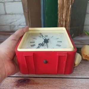 May include: A vintage red and cream-colored alarm clock with a square face. The clock has a white face with black Roman numerals and hands. The clock is held in a hand.