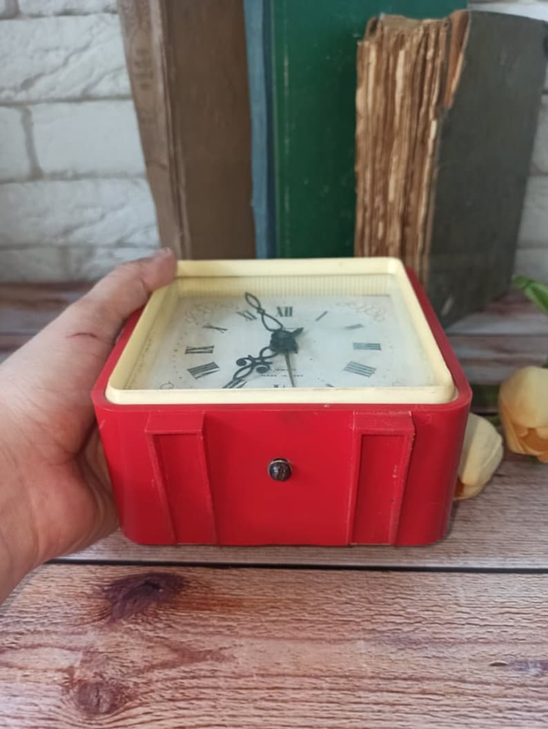 May include: A vintage red and cream-colored alarm clock with a square face. The clock has a white face with black Roman numerals and hands. The clock is held in a hand.