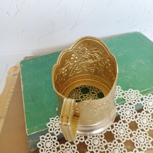 May include: Gold metal cup holder with an ornate design. The holder has a handle and is sitting on a book and a doily.