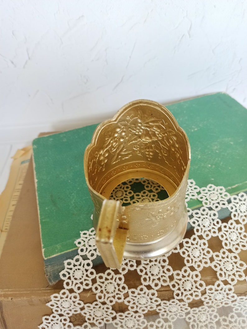 May include: Gold metal cup holder with an ornate design. The holder has a handle and is sitting on a book and a doily.