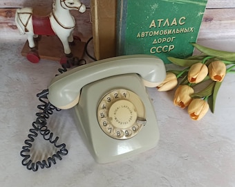 May include: A vintage rotary dial phone with a green body and a white dial. The dial has black numbers and the text "RIGA - LATVIJAS TELEFONS" around the edge. The phone has a black coiled cord.