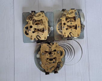 May include: Three vintage clock movements with brass gears and metal plates. The clock mechanisms are arranged in a triangular pattern, showcasing the intricate internal workings of the timepieces. A coiled spring is also visible.
