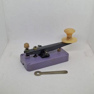 May include: A vintage purple telegraph key with a black lever and a white plastic knob. The key is on a white surface with a small metal wrench.