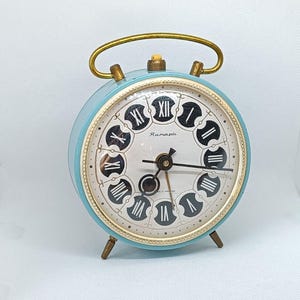 May include: A light blue vintage alarm clock with a white face and black Roman numeral hour markers. The clock has a gold-colored handle and legs. The clock face says "Rexmapi".
