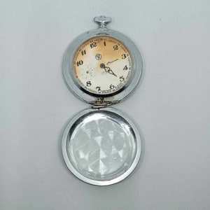 Può includere: Un orologio da tasca argentato con un quadrante bianco e numeri neri. L'orologio ha una piccola lancetta dei secondi e una lancetta dei minuti più grande. L'orologio è aperto e il retro è visibile. Il retro è anche argentato e ha un motivo decorativo.