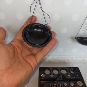 Puede incluir: Una balanza de equilibrio vintage negra con un cuenco negro colgando de un brazo y un juego de pesas en una caja negra. La balanza es de metal y tiene una cuerda azul.