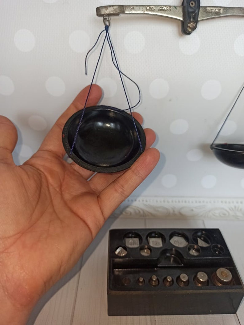 Puede incluir: Una balanza de equilibrio vintage negra con un cuenco negro colgando de un brazo y un juego de pesas en una caja negra. La balanza es de metal y tiene una cuerda azul.