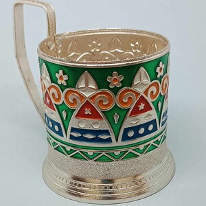 May include: A silver-colored metal cup holder with a green, orange, white, and blue geometric design. The cup holder has a handle and a textured base.