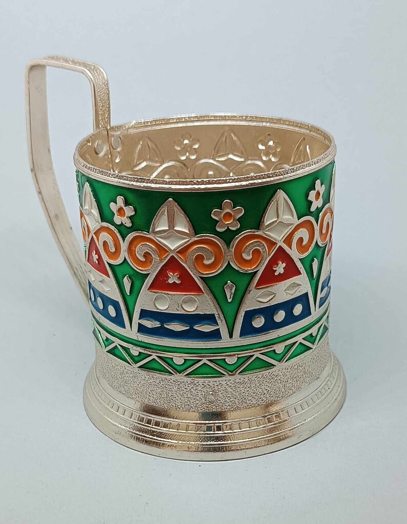 May include: A silver-colored metal cup holder with a green, orange, white, and blue geometric design. The cup holder has a handle and a textured base.