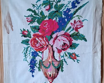 May include: A vintage needlepoint wall hanging featuring a bouquet of pink, red, and blue flowers in a green and white vase. The flowers are arranged in a symmetrical pattern and are surrounded by green leaves. The background is a white fabric with a subtle texture.