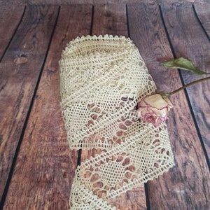 May include: A close-up of a beige crocheted lace trim on a wooden background. A dried rose is partially visible in the lower right corner of the image.
