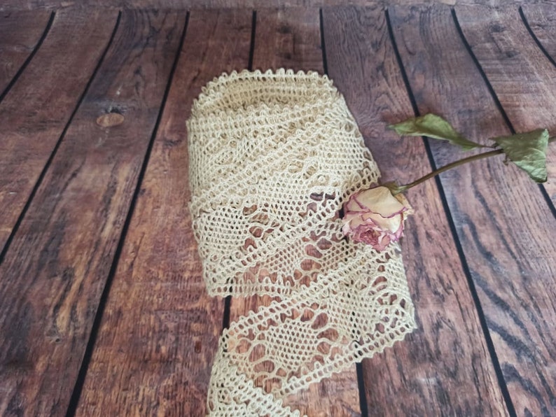 May include: A close-up of a beige crocheted lace trim on a wooden background. A dried rose is partially visible in the lower right corner of the image.