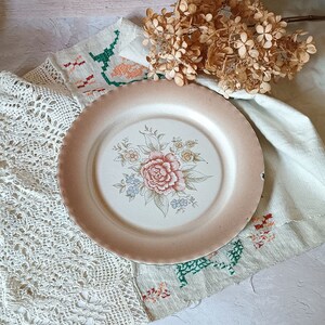 Puede incluir: Un plato vintage con diseño floral, fondo crema y un patrón floral rosa, blanco y verde. El plato está sobre un mantel de crochet blanco con un arreglo de flores secas.