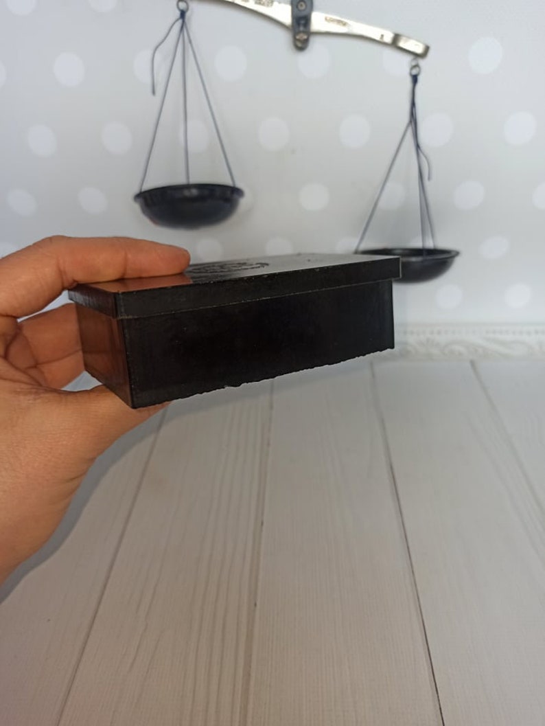 Puede incluir: Una caja rectangular negra con una tapa ligeramente elevada. La caja est&aacute; hecha de un material liso y duro y tiene un brillo sutil. La caja est&aacute; siendo sostenida en una mano, pero la mano no es visible en la imagen.